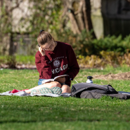 Student wearing a University of Chicago sweatshirt reading a book while sitting on a blanket on the grass.